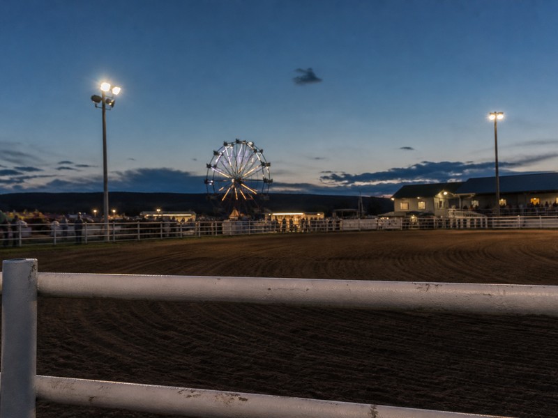 A county fair Ferris wheel glowing beyond an empty rodeo arena at dusk.